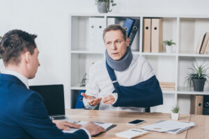 middle aged worker in neck brace with broken arm sitting at table and talking to businessman in blue jacket in office