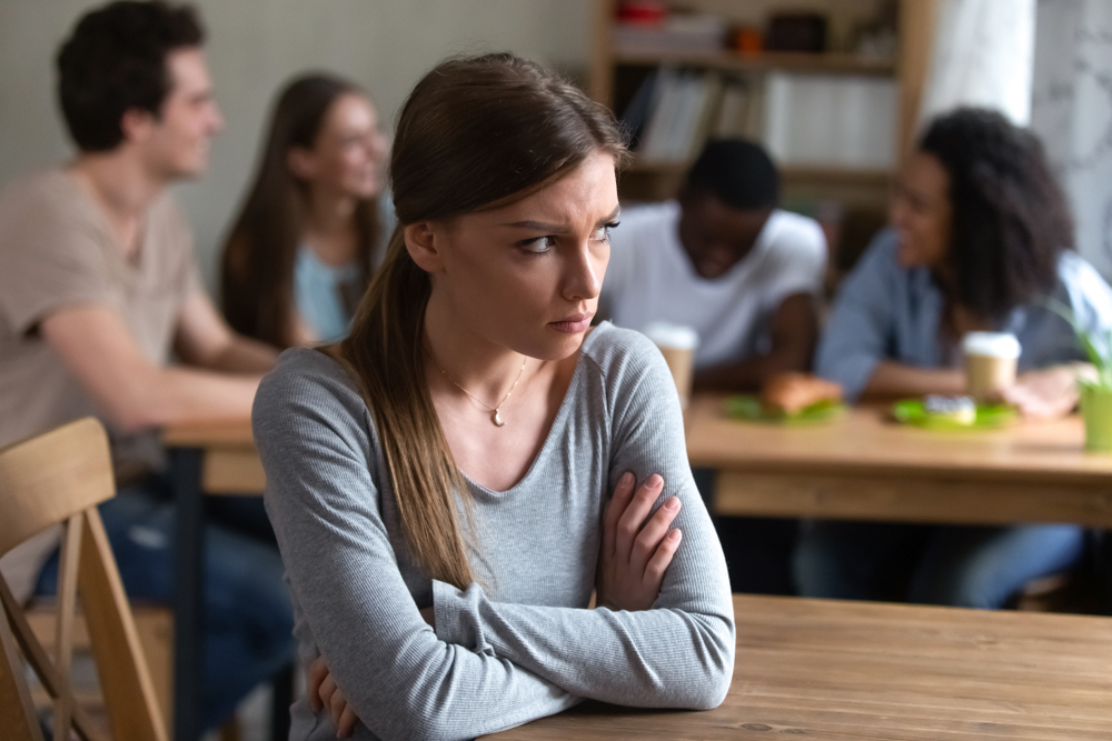 Abused upset offended by friends young caucasian woman sitting alone separately from group of happy laughing people